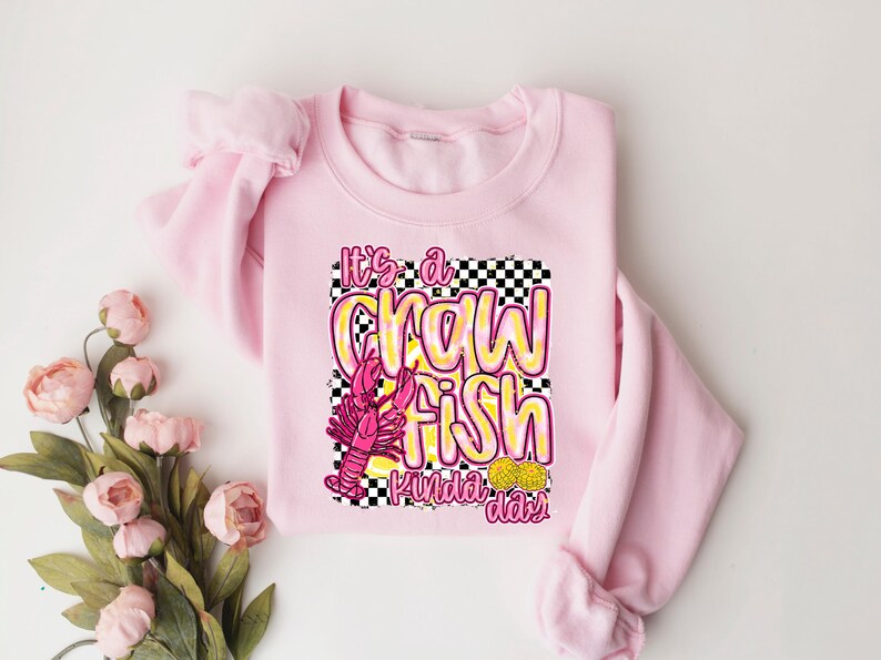 Puede incluir: Sudadera rosa con un dise&ntilde;o gr&aacute;fico de un cangrejo de r&iacute;o y el texto "It's a crawfish kinda day".