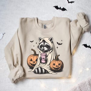 Puede incluir: Una sudadera de color beige con un gr&aacute;fico de un mapache vestido con un disfraz de fantasma y sosteniendo una bebida rosa. El mapache est&aacute; rodeado de tres calabazas con caras de calabaza.