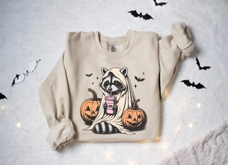 Puede incluir: Una sudadera de color beige con un gr&aacute;fico de un mapache vestido con un disfraz de fantasma y sosteniendo una bebida rosa. El mapache est&aacute; rodeado de tres calabazas con caras de calabaza.