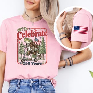 May include: Pink t-shirt with a graphic featuring a frog riding a scooter, holding an American flag, and the text "Off To Celebrate 250 Years." An American flag patch is on the sleeve.