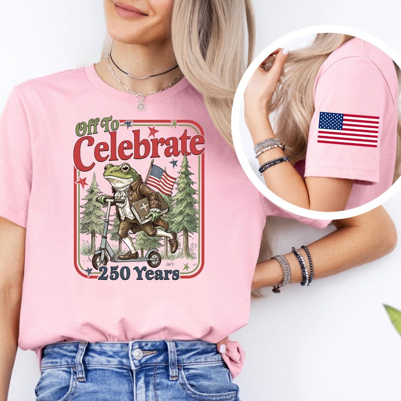 May include: Pink t-shirt with a graphic featuring a frog riding a scooter, holding an American flag, and the text "Off To Celebrate 250 Years." An American flag patch is on the sleeve.