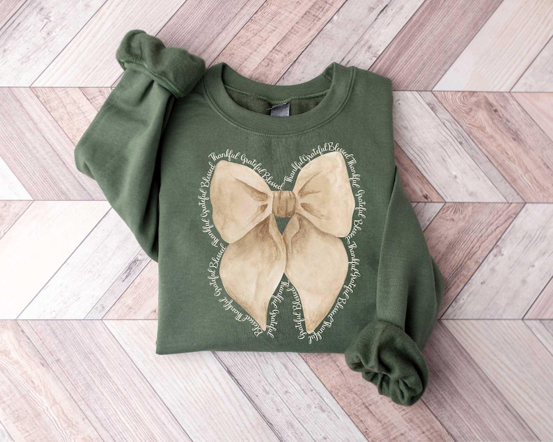 May include: A green sweatshirt with a large brown bow on the front. The bow is surrounded by the words "Thankful Grateful Blessed" repeated around the bow.