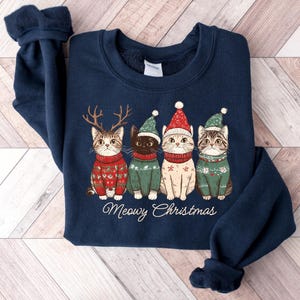 May include: A navy blue sweatshirt with a Christmas-themed design featuring four cats wearing Santa hats and sweaters. The text "Meowy Christmas" is printed below the cats.