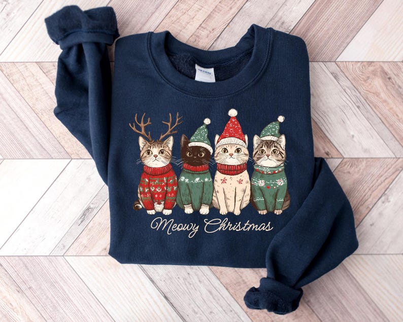 May include: A navy blue sweatshirt with a Christmas-themed design featuring four cats wearing Santa hats and sweaters. The text "Meowy Christmas" is printed below the cats.
