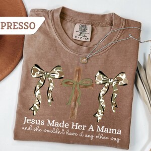 May include: Espresso-colored t-shirt with a graphic design featuring a cross and two bows in a camouflage pattern. The text "Jesus Made Her A Mama and she wouldn't have it any other way" is printed below the design. A silver necklace is draped across the shirt.