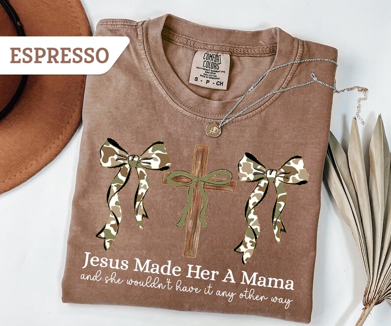 May include: Espresso-colored t-shirt with a graphic design featuring a cross and two bows in a camouflage pattern. The text "Jesus Made Her A Mama and she wouldn't have it any other way" is printed below the design. A silver necklace is draped across the shirt.