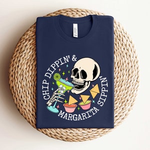 Pu&ograve; includere: Una maglietta blu navy con una grafica bianca di uno scheletro che beve una margarita e intinge patatine. Il testo sulla maglietta recita "Chip Dippin' & Margarita Sippin'."