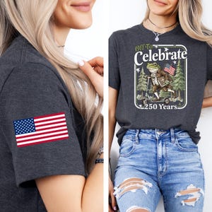 May include: Dark gray t-shirts with an American flag patch on the sleeve and a graphic of a frog riding a scooter with the text "Off To Celebrate 250 Years." The frog is holding an American flag and a book.