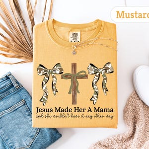 May include: Mustard-colored t-shirt with a graphic featuring a cross, two bows with camouflage print, and the text "Jesus Made Her A Mama and she wouldn't have it any other way." A gold necklace is visible. The shirt is folded on a white surface.