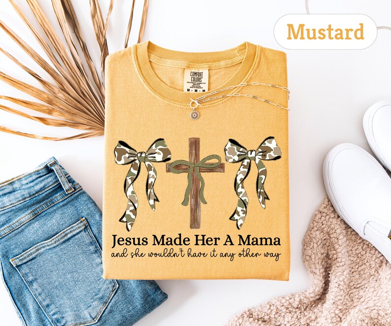 May include: Mustard-colored t-shirt with a graphic featuring a cross, two bows with camouflage print, and the text "Jesus Made Her A Mama and she wouldn't have it any other way." A gold necklace is visible. The shirt is folded on a white surface.