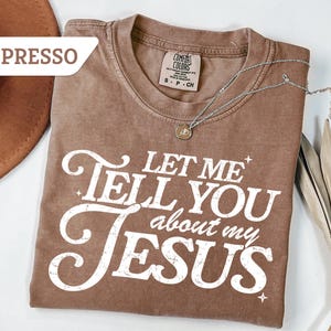 Puede incluir: Camiseta color espresso con el texto blanco "LET ME TELL YOU about my JESUS". La camiseta tiene un aspecto vintage y desgastado. Un collar plateado con un colgante est&aacute; drapeado sobre la camiseta. Un sombrero marr&oacute;n es visible en el fondo.