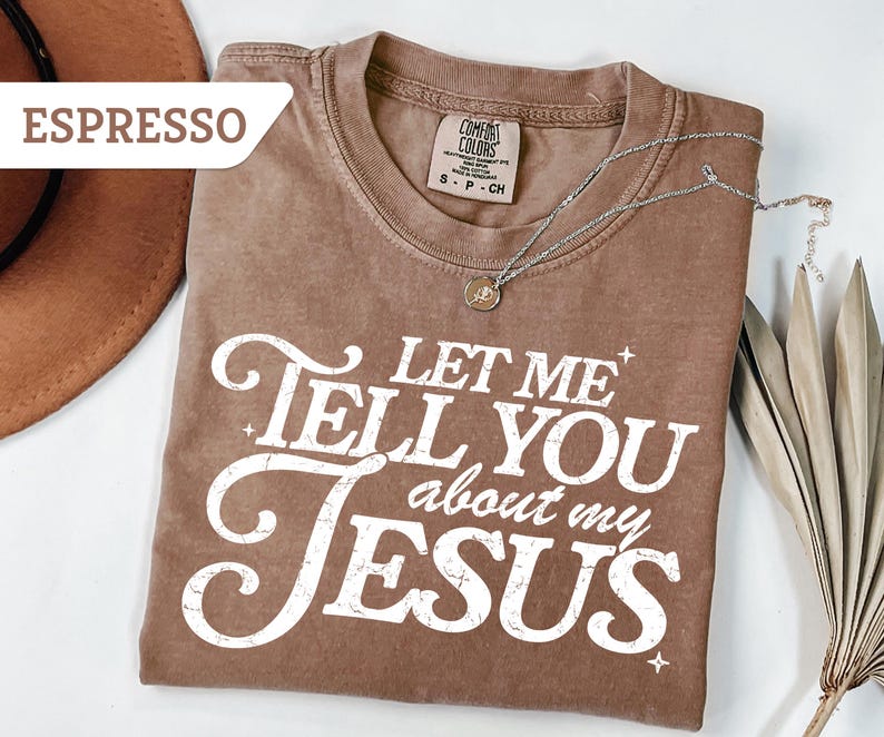 Puede incluir: Camiseta color espresso con el texto blanco "LET ME TELL YOU about my JESUS". La camiseta tiene un aspecto vintage y desgastado. Un collar plateado con un colgante est&aacute; drapeado sobre la camiseta. Un sombrero marr&oacute;n es visible en el fondo.