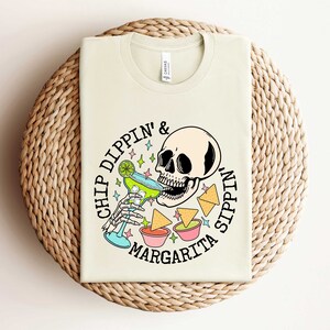 Pu&ograve; includere: Una maglietta beige con una grafica di uno scheletro che beve una margarita e il testo "Chip Dippin' & Sippin' Margarita".
