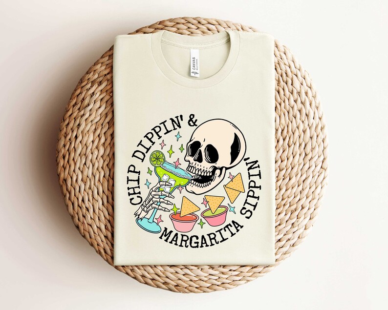 Pu&ograve; includere: Una maglietta beige con una grafica di uno scheletro che beve una margarita e il testo "Chip Dippin' & Sippin' Margarita".
