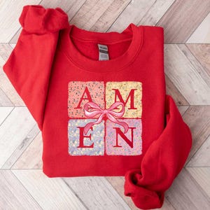 K&ouml;nnte beinhalten: Roter Pullover mit einem Aufdruck einer Geschenkbox mit dem Wort "AMEN" in floral gemusterten Quadraten. Die Geschenkbox hat eine rosafarbene Schleife.