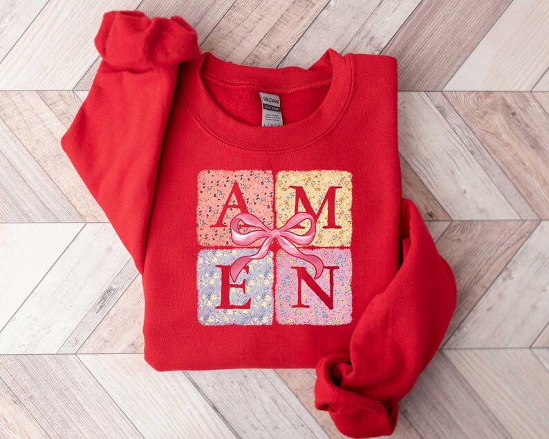 K&ouml;nnte beinhalten: Roter Pullover mit einem Aufdruck einer Geschenkbox mit dem Wort "AMEN" in floral gemusterten Quadraten. Die Geschenkbox hat eine rosafarbene Schleife.