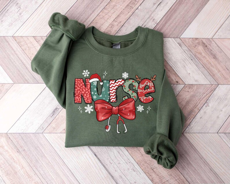 May include: Green sweatshirt with a Christmas-themed "Nurse" graphic. The letters are decorated with snowflakes, holly, and a Santa hat. A red bow with a stethoscope is below the word.