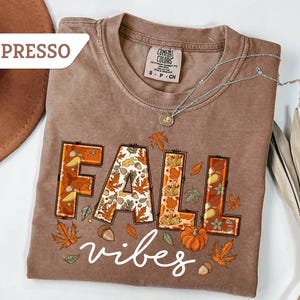 May include: Espresso-colored t-shirt with the words "FALL vibes" in a fall-themed design. The word "FALL" is in a block letter style with autumn leaves and acorns. A pumpkin and leaves are also featured.