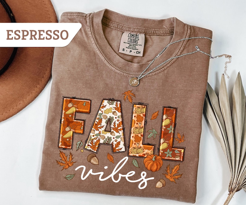 May include: Espresso-colored t-shirt with the words "FALL vibes" in a fall-themed design. The word "FALL" is in a block letter style with autumn leaves and acorns. A pumpkin and leaves are also featured.