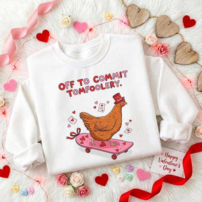 K&ouml;nnte beinhalten: Wei&szlig;es Sweatshirt mit dem Text "OFF TO COMMIT TOMFOOLERY." Ein Cartoon-Huhn mit Zylinder f&auml;hrt auf einem rosa Skateboard mit Herzen. Das Design enth&auml;lt Herzen und Valentinstag-Elemente.