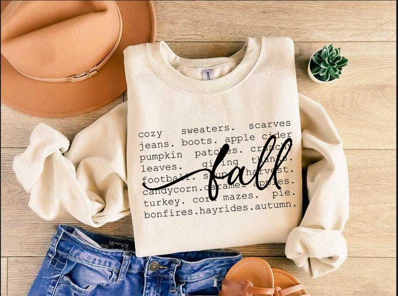 Pu&ograve; includere: Una felpa beige con la scritta "cozy sweaters. scarves jeans. boots. apple cider pumpkin patches. crackling leaves. giving thanks. fall football. soup harvest. candycorn. caramel apples. turkey. corn mazes. pie. bonfires. hayrides. autumn." stampata in nero.