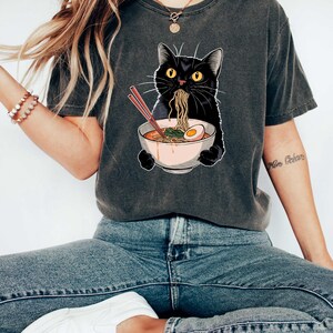 May include: A black cat wearing a grey t-shirt with a graphic of a black cat eating ramen noodles. The cat is holding a bowl of ramen with chopsticks.
