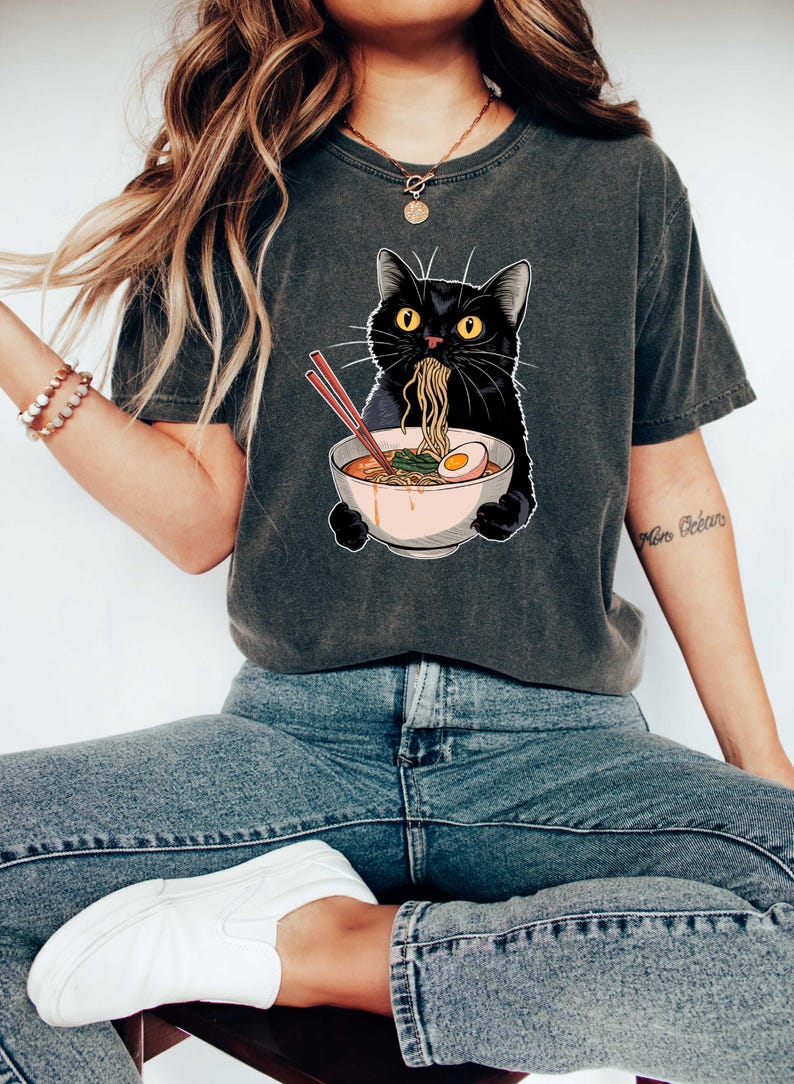 May include: A black cat wearing a grey t-shirt with a graphic of a black cat eating ramen noodles. The cat is holding a bowl of ramen with chopsticks.