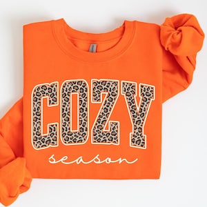 May include: An orange sweatshirt with the words "COZY" and "season" printed on the front. The word "COZY" is in a bold, block letter style with a leopard print pattern. The word "season" is written in a cursive font.