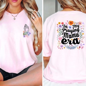May include: Pink t-shirt with a floral design and the text "In my Praying Mama era" on the back. The front of the shirt has a small design of hands in a praying position with flowers.