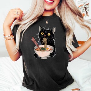 May include: Black t-shirt with a graphic of a black cat eating ramen noodles from a bowl with chopsticks. The cat has a surprised expression.
