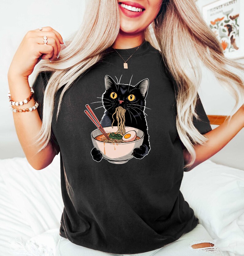 May include: Black t-shirt with a graphic of a black cat eating ramen noodles from a bowl with chopsticks. The cat has a surprised expression.