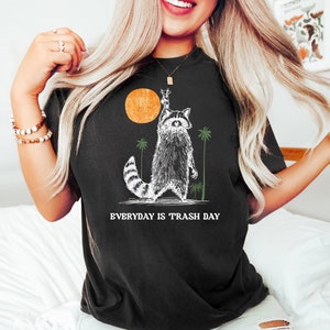 Może przedstawiać: Czarny T-shirt z grafiką przedstawiającą szopa pracza podnoszącego znak pokoju przed zachodem słońca. Pod szopem praczem nadrukowany jest tekst "EVERYDAY IS TRASH DAY".
