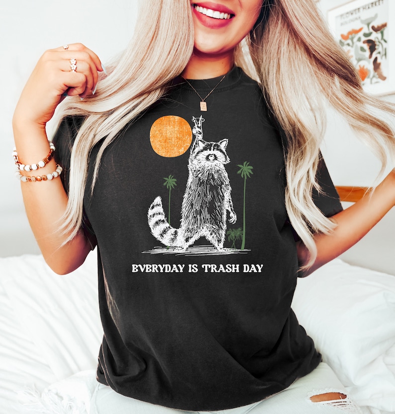 Może przedstawiać: Czarny T-shirt z grafiką przedstawiającą szopa pracza podnoszącego znak pokoju przed zachodem słońca. Pod szopem praczem nadrukowany jest tekst "EVERYDAY IS TRASH DAY".