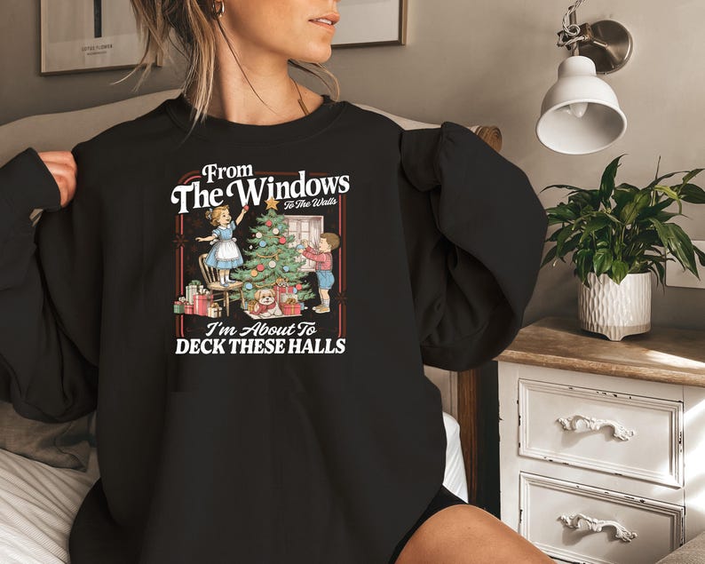 May include: Black sweatshirt with a Christmas-themed graphic. The graphic features a decorated Christmas tree, presents, and two children. Text on the sweatshirt reads "From The Windows To The Walls, I'm About To DECK THESE HALLS."