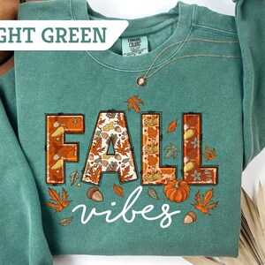 May include: Light green sweatshirt with the words "FALL vibes" in a fall-themed design. The letters are outlined in brown and filled with autumn leaves, acorns, and a pumpkin. The sweatshirt has a crew neck and long sleeves.