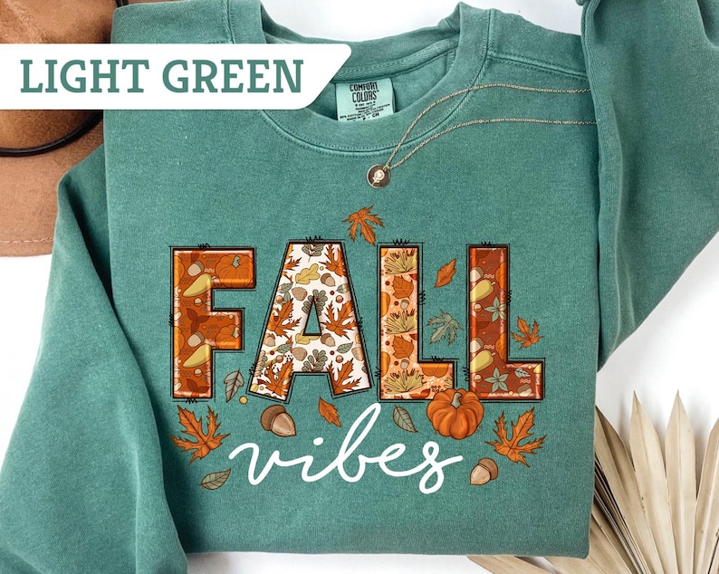 May include: Light green sweatshirt with the words "FALL vibes" in a fall-themed design. The letters are outlined in brown and filled with autumn leaves, acorns, and a pumpkin. The sweatshirt has a crew neck and long sleeves.