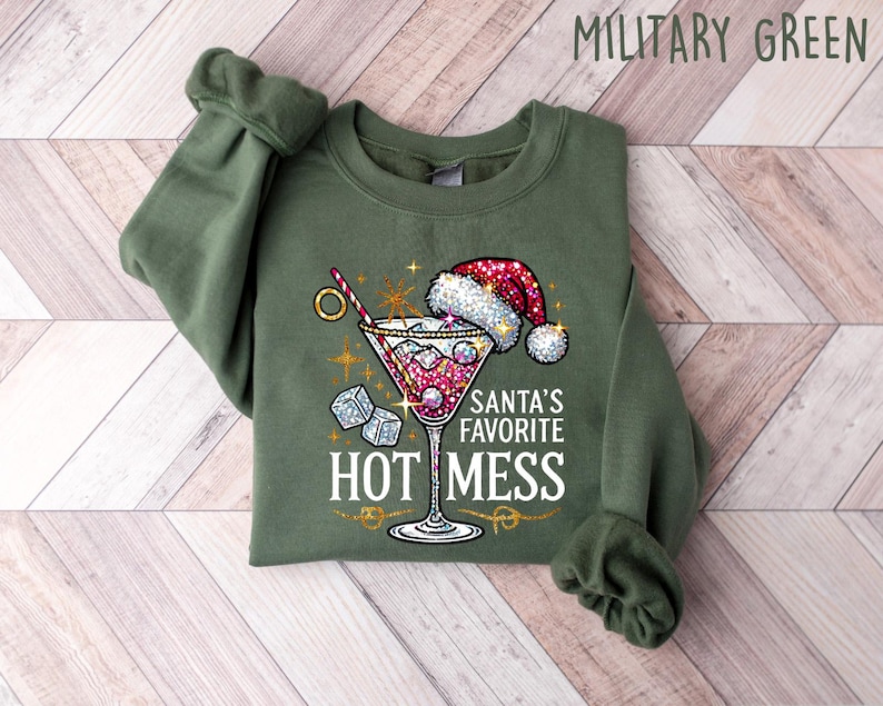 May include: Military green Christmas sweatshirt featuring a sparkly martini glass graphic with a Santa hat on the rim. Text on the sweatshirt reads, 'Santa's Favorite Hot Mess.' The sweatshirt is a crewneck style.
