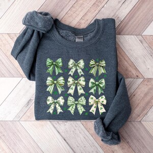 May include: A dark gray sweatshirt with a design of 12 green and white bows with shamrock accents. The bows are arranged in a grid pattern.