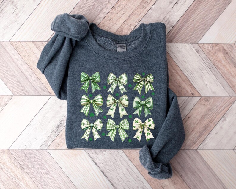 May include: A dark gray sweatshirt with a design of 12 green and white bows with shamrock accents. The bows are arranged in a grid pattern.