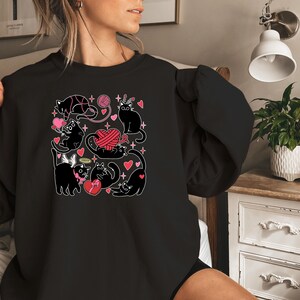 May include: Black sweatshirt with a graphic print of black cats playing with yarn and hearts. The cats are wearing angel wings and a crown.