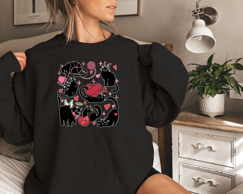 May include: Black sweatshirt with a graphic print of black cats playing with yarn and hearts. The cats are wearing angel wings and a crown.