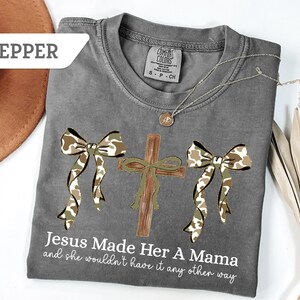 May include: A gray Comfort Colors t-shirt with a cross and two bows in a camouflage pattern. The text "Jesus Made Her A Mama and she wouldn't have it any other way" is printed on the shirt. A silver necklace and a brown hat are also visible.