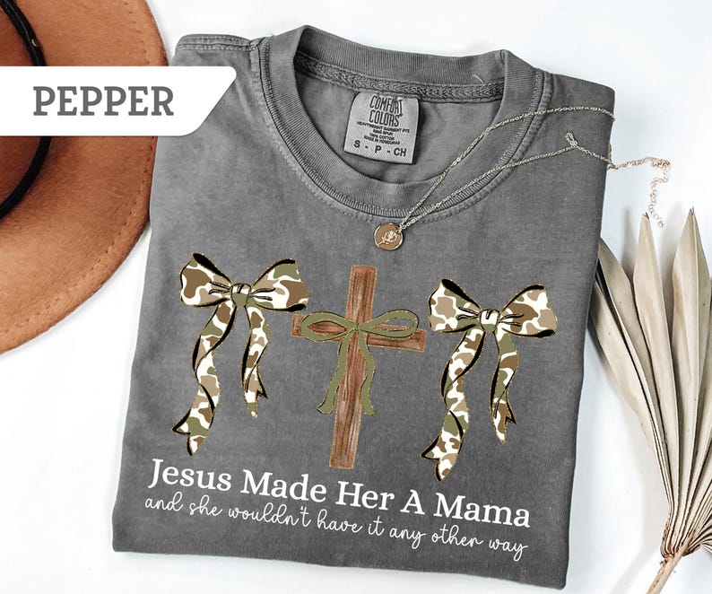 May include: A gray Comfort Colors t-shirt with a cross and two bows in a camouflage pattern. The text "Jesus Made Her A Mama and she wouldn't have it any other way" is printed on the shirt. A silver necklace and a brown hat are also visible.