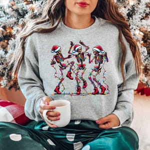 May include: A gray sweatshirt with a festive design of four skeletons wearing Santa hats and dancing with Christmas lights.