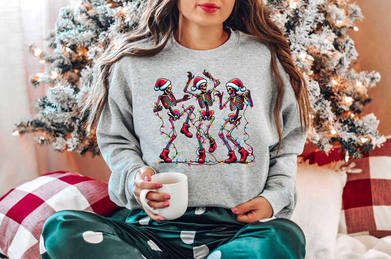 May include: A gray sweatshirt with a festive design of four skeletons wearing Santa hats and dancing with Christmas lights.