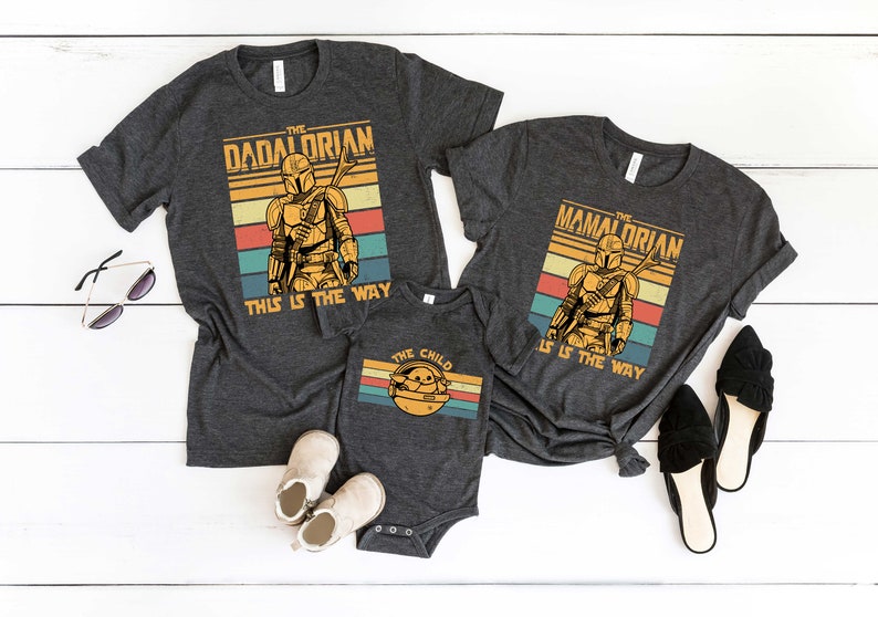 May include: Three gray t-shirts with a retro design featuring a Mandalorian character. The shirts have the text "The Dadalorian", "The Mandalorian", and "The Child" with the phrase "This is the way" on the larger shirts.