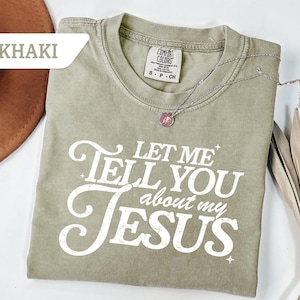 Puede incluir: Camiseta caqui con el texto blanco "LET ME TELL YOU about my JESUS". Un collar plateado con un colgante descansa sobre la camiseta. La camiseta es de la marca Comfort Colors.