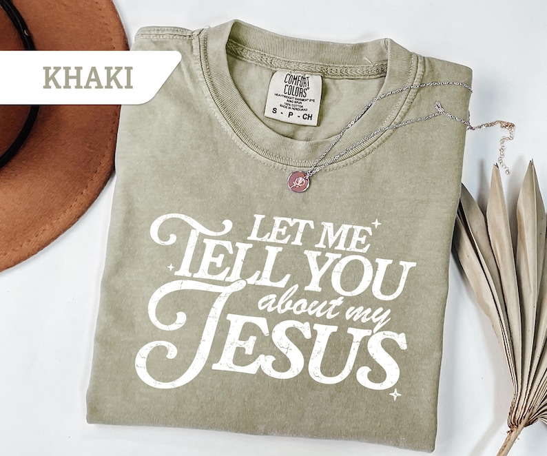 Puede incluir: Camiseta caqui con el texto blanco "LET ME TELL YOU about my JESUS". Un collar plateado con un colgante descansa sobre la camiseta. La camiseta es de la marca Comfort Colors.