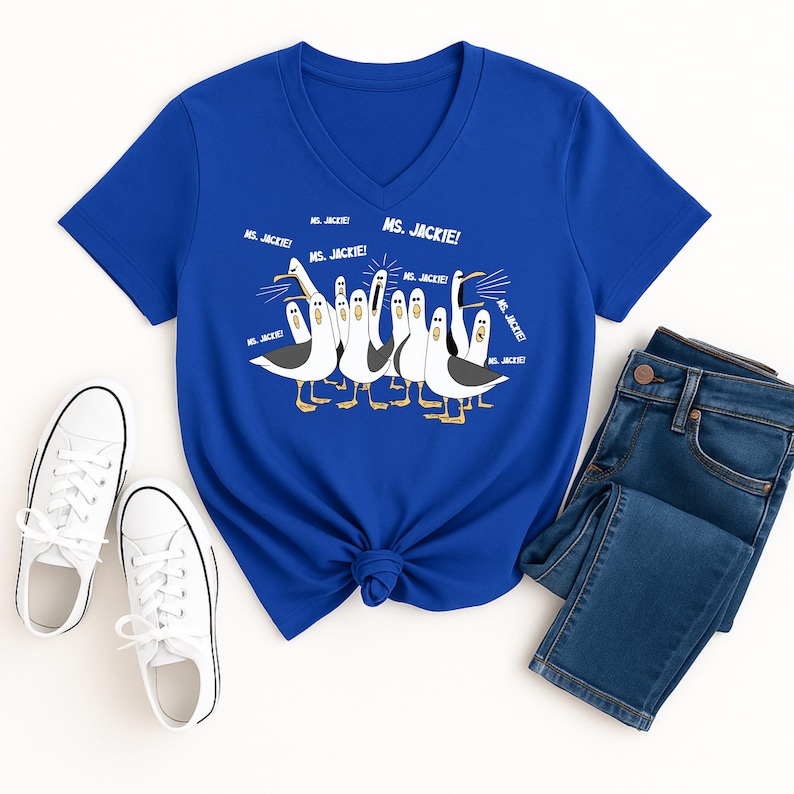 May include: A royal blue V-neck t-shirt featuring a cartoon illustration of seagulls with the text "MS. JACKIE!". The shirt is paired with blue jeans and white sneakers.