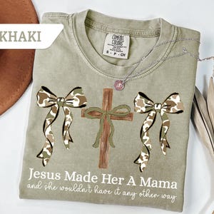 May include: Khaki-colored t-shirt with a cross and two camo bows. The text "Jesus Made Her A Mama and she wouldn't have it any other way" is printed on the shirt. A silver necklace with a pendant is on the shirt.